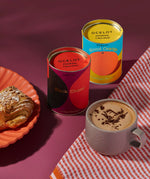 Ocelot Black Cherry and blood orange Organic Drinking Chocolate tins placed next to a cup of hot chocolate and a pasty on a plate.
