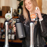 Woman holding a One Green Bottle plastic free stainless steel water bottle next to some beer pumps 