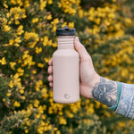 Close up of a hand holding a 500ml One Green Bottle drinks canteen in front of some yellow and green bushes