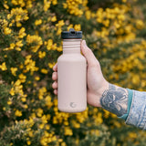 Close up of a hand holding a 500ml One Green Bottle drinks canteen in front of some yellow and green bushes