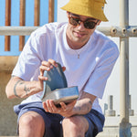 A man sitting on a stone surface and lifting the pale blue silicone lid from the soldier One Green Bottle lunchbox.
