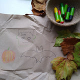 OkoNorm 6 stubby beeswax crayons placed into a jute basket next to Halloween themed drawings on brown paper. 
