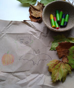 OkoNorm 6 stubby beeswax crayons placed into a jute basket next to Halloween themed drawings on brown paper. 
