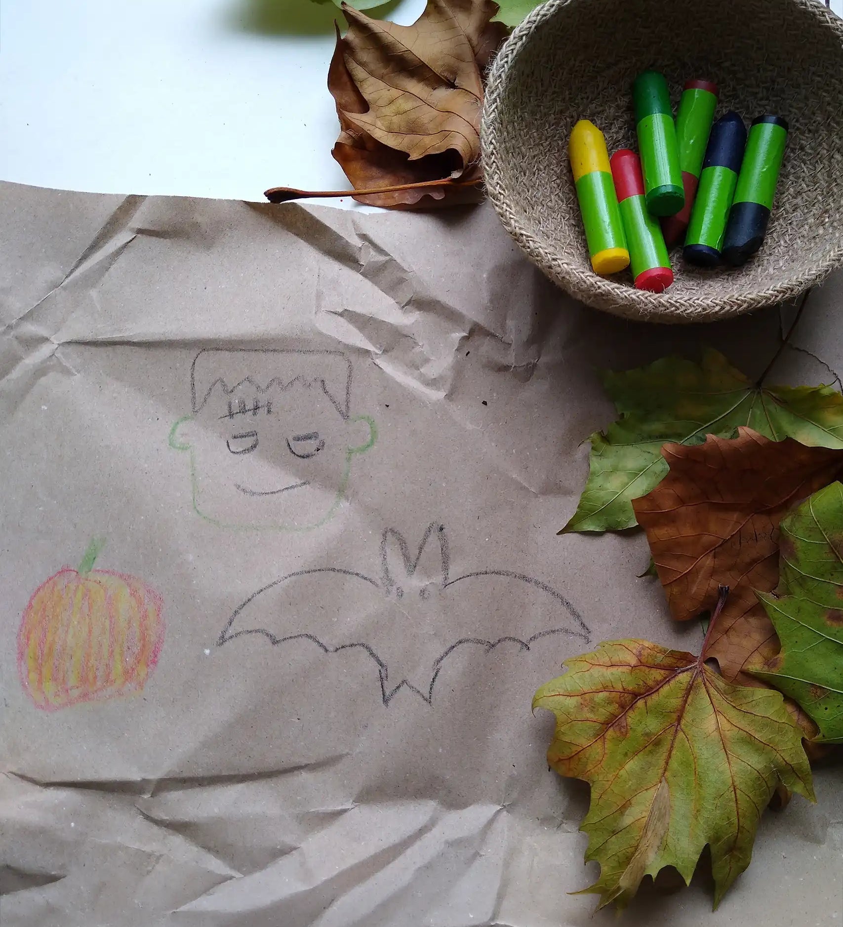 OkoNorm 6 stubby beeswax crayons placed into a jute basket next to Halloween themed drawings on brown paper. 
