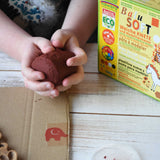 A close up of a child's hands holding a handful of brown colour Okonorm play dough from the earth colour set. The cardboard packaging box that the set of four play dough comes in is next to the child. 
