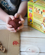 A close up of a child's hands holding a handful of brown colour Okonorm play dough from the earth colour set. The cardboard packaging box that the set of four play dough comes in is next to the child. 