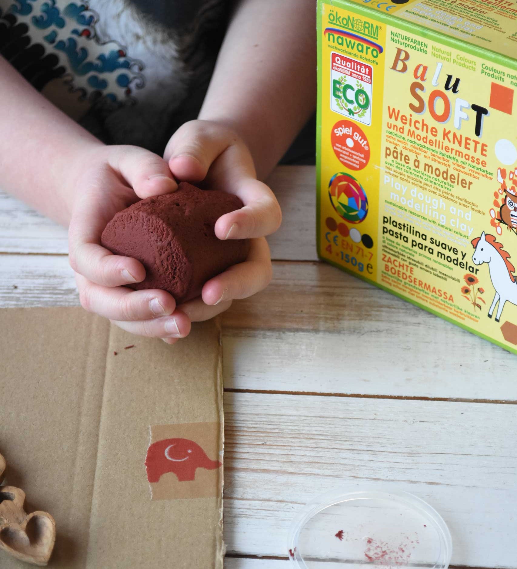 A close up of a child's hands holding a handful of brown colour Okonorm play dough from the earth colour set. The cardboard packaging box that the set of four play dough comes in is next to the child. 
