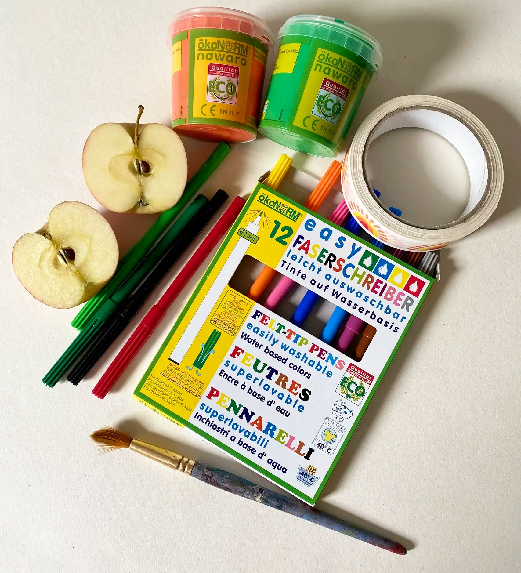 Pack of OkoNorm 12 felt tip pens placed besides Okonorm paints, Babipur tapes and a cut apple.