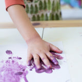Child's hand covered in purple Okonorm finger paints placing it on a piece of paper to make a handprint.