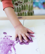 Child's hand covered in purple Okonorm finger paints placing it on a piece of paper to make a handprint.