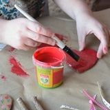 A red pot of Okonorm finger paint placed on some brown paper. A child can be seen painting a heart shaped piece of cardboard in the background.