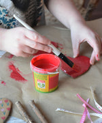 A red pot of Okonorm finger paint placed on some brown paper. A child can be seen painting a heart shaped piece of cardboard in the background.