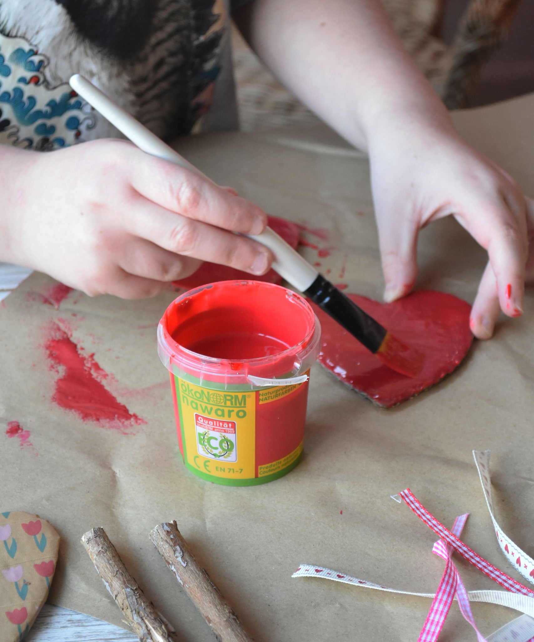 A red pot of Okonorm finger paint placed on some brown paper. A child can be seen painting a heart shaped piece of cardboard in the background.