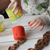 Child tipping red Okonorm play dough out of the storage tub onto a wooden surface next to Welsh love spoons.