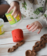 Child tipping red Okonorm play dough out of the storage tub onto a wooden surface next to Welsh love spoons.