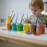 OkoNorm finger paints in 6 unicorn  colours. Red, orange, yellow, green blue and purple pots lined up next to each other with the lids off. Child painting in the background.