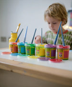 OkoNorm finger paints in 6 unicorn  colours. Red, orange, yellow, green blue and purple pots lined up next to each other with the lids off. Child painting in the background.