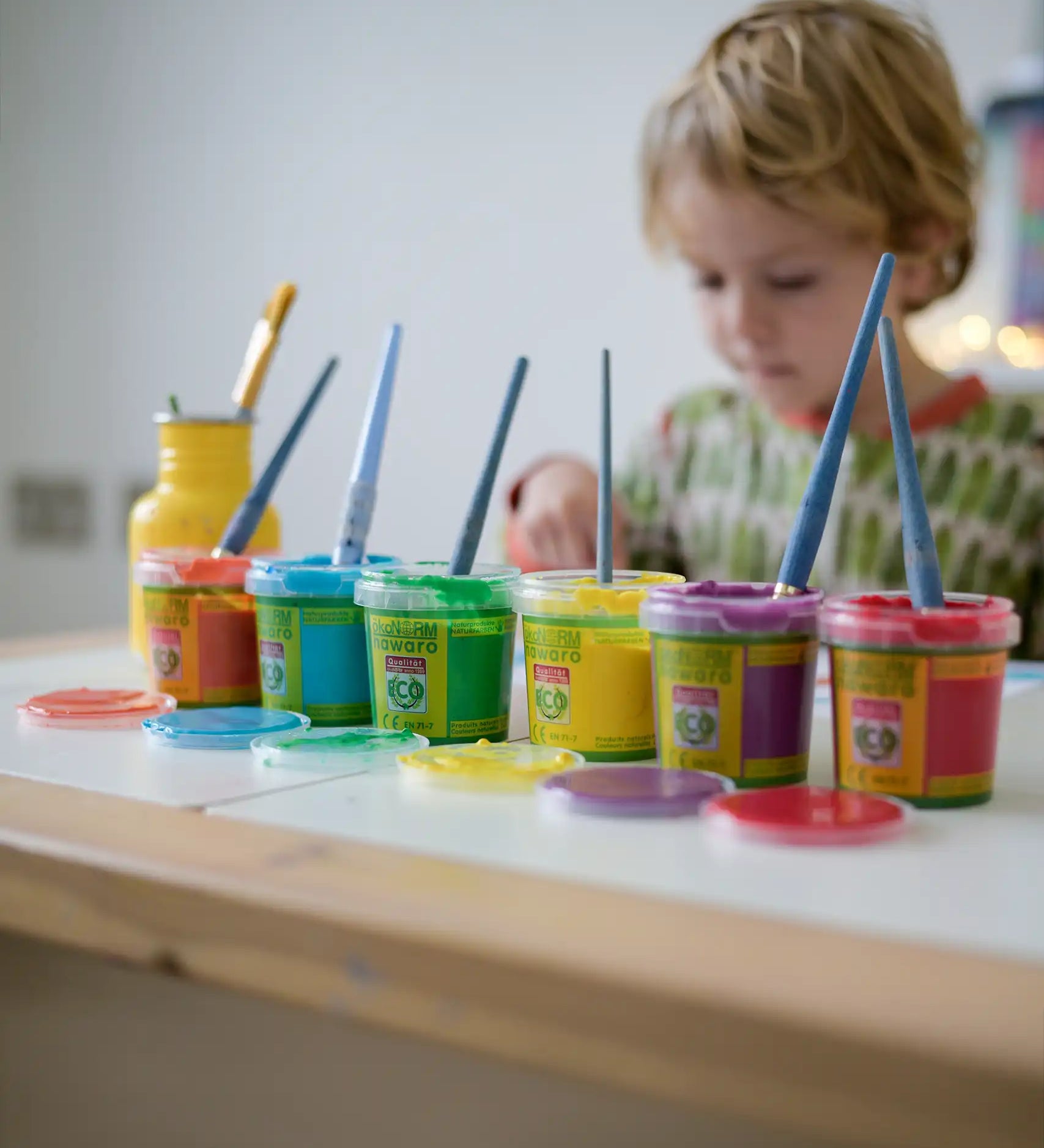 OkoNorm finger paints in 6 unicorn  colours. Red, orange, yellow, green blue and purple pots lined up next to each other with the lids off. Child painting in the background.