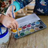 Children placing paintbrushes on the watercolour pods from the Okonorm 12 colour watercolour paints in a travel tin.