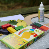 Okonorm 12 colour wax blocks pack placed on a table outdoors with other craft supplies and okonorm all purpose craft glue. 