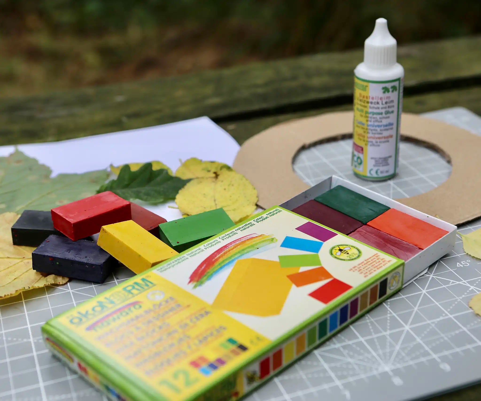 Okonorm 12 colour wax blocks pack placed on a table outdoors with other craft supplies and okonorm all purpose craft glue. 