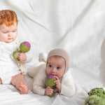 One baby sitting holding an Oli & Carol Artichoke Rattle Teether toy with another baby lying by their side chewing on an Oli & Carol Artichoke Rattle Teether