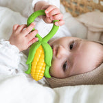 baby lying down in a changing basket clutching onto the Oli & Carol Corn Rattle Teether
