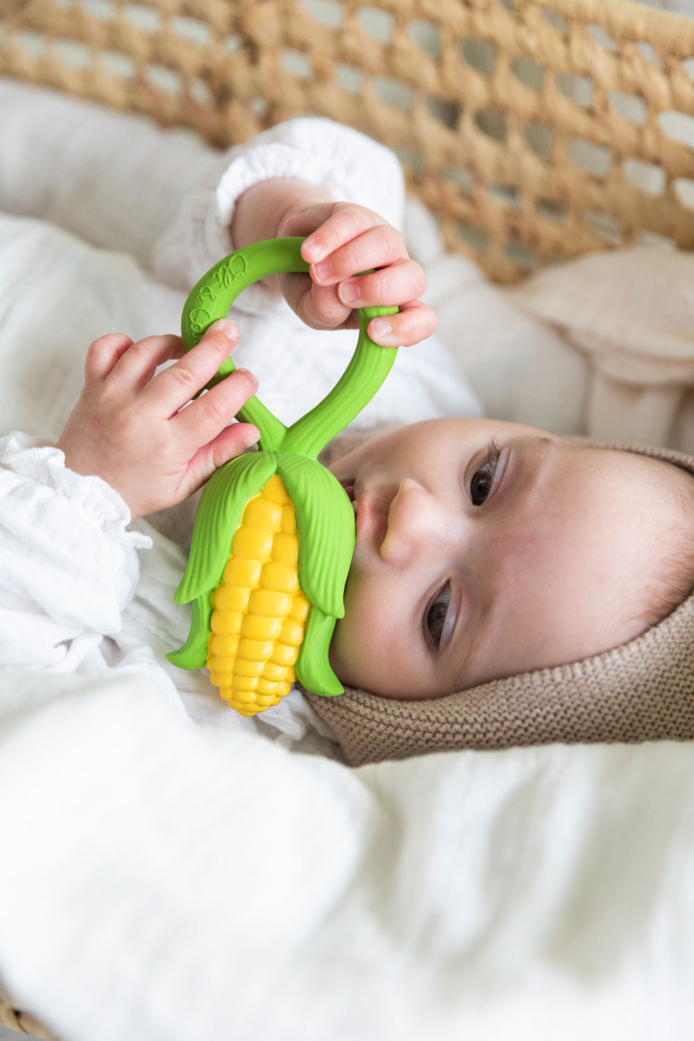 baby lying down in a changing basket clutching onto the Oli & Carol Corn Rattle Teether