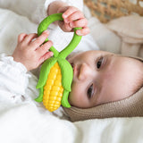 baby lying down in a changing basket clutching onto the Oli & Carol Corn Rattle Teether