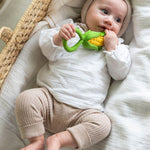 baby lying down in a changing basket clutching onto the Oli & Carol Corn Rattle Teether