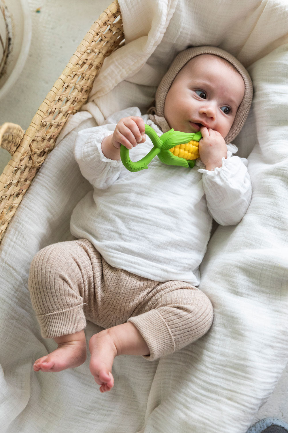 baby lying down in a changing basket clutching onto the Oli & Carol Corn Rattle Teether