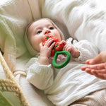 Baby chewing the Oli & Carol Tomato Rattle Teether lying down on a white muslin cloth