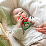Baby chewing the Oli & Carol Tomato Rattle Teether lying down on a white muslin cloth