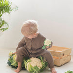 A child sitting down holding an Oli & Carol Cauliflower Rattle Teether with real cauliflowers in front of them