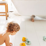 Child in bathroom playing with yellow and mint coloured Oli & Carol Flo The Floatie Ducks natural rubber bath toys