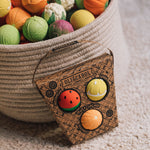 A packet of Oli & Carol Fruit Salad Baby Sensory Balls including a watermelon, lemon and tangerine in front of a basket  full of Oli & Carol Baby Sensory balls.