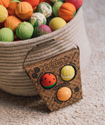 A packet of Oli & Carol Fruit Salad Baby Sensory Balls including a watermelon, lemon and tangerine in front of a basket  full of Oli & Carol Baby Sensory balls.