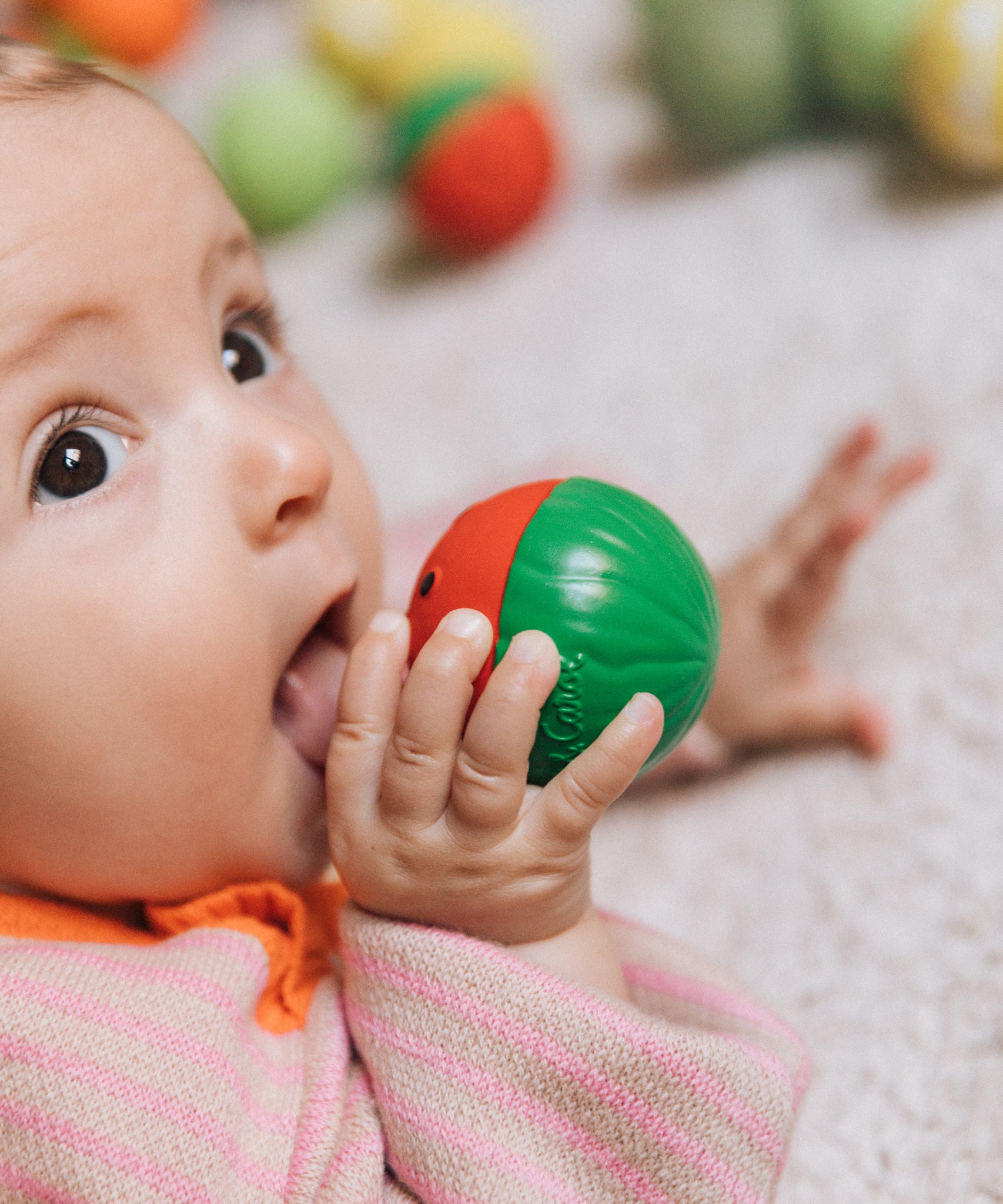 A baby holding onto a watermelon Oli & Carol sensory ball near their mouth