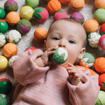 A baby clutching onto a cauliflower ball from the Oli & Carol Vegetable soup Baby Sensory Balls set surrounded by fruit and vegetable sensory balls.