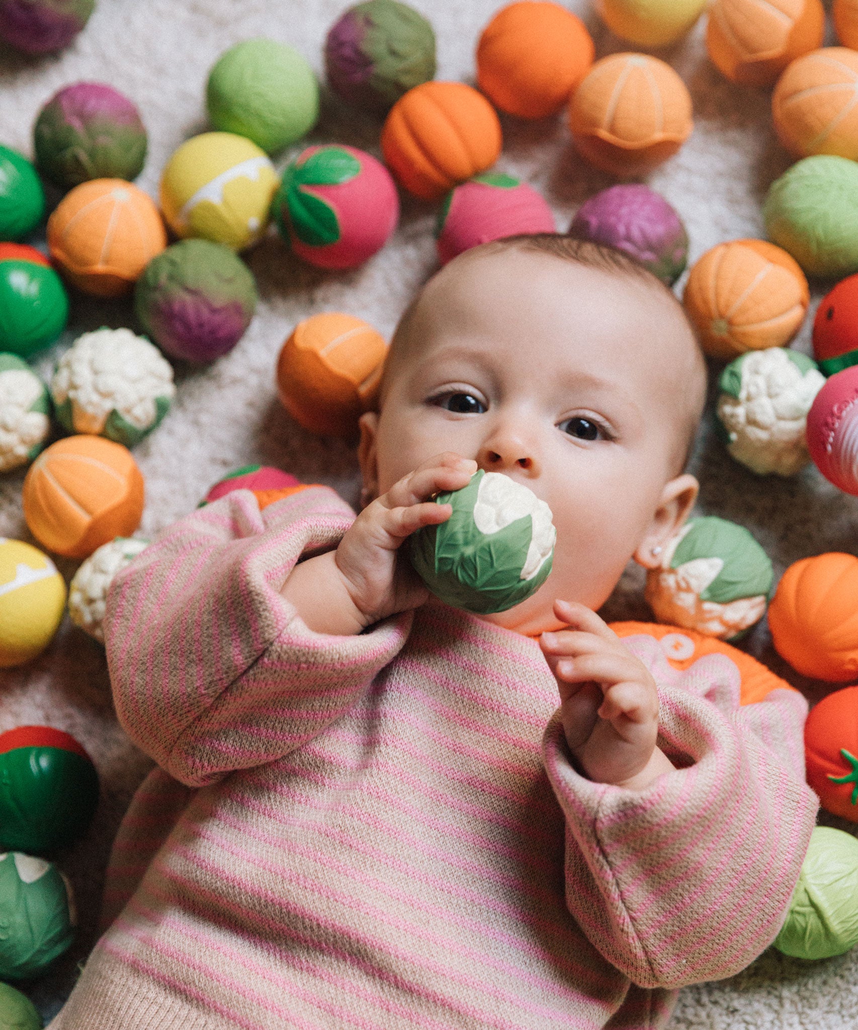 A baby clutching onto a cauliflower ball from the Oli & Carol Vegetable soup Baby Sensory Balls set surrounded by fruit and vegetable sensory balls.