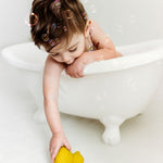 Baby in a small bath tub holding reaching for a yellow Oli & Carol Elvis The Duck Bath Toy which is on the floor