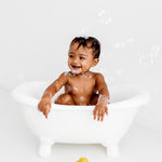 Baby in a small bath tub holding with bubbles floating around with a yellow Oli & Carol Elvis The Duck Bath Toy on the floor in front