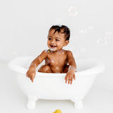 Baby in a small bath tub holding with bubbles floating around with a yellow Oli & Carol Elvis The Duck Bath Toy on the floor in front