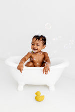 Baby in a small bath tub holding with bubbles floating around with a yellow Oli & Carol Elvis The Duck Bath Toy on the floor in front