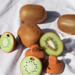 Oli & Carol Jose Antonio The Kiwi Mini Baby Teethers pictured amongst real kiwi fruit placed on a white cotton sheet