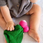 Close up of the Oli & Carol eco-friendly mini radish baby comforter toy on a grey sheet next to a baby's leg
