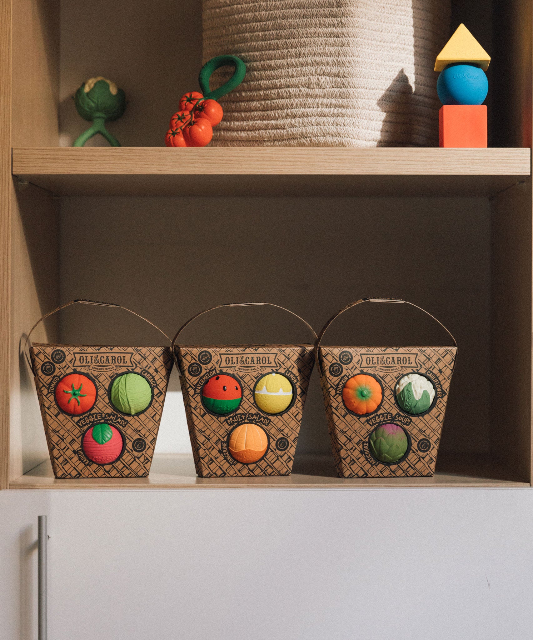 Three Packets of Oli & Carol Baby Sensory Balls, Fruit Salad, Veggie Salad and Veggie Soup placed on shelving with other Oli and Carol toys above them