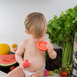 Child chewing on teh Oli & Carol Renato The Tomato with a pile of other Oli & Carol toys by their side