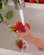 Close up of a childs hand holding the oli and carol soft rubber raspberry food toy under a running tap