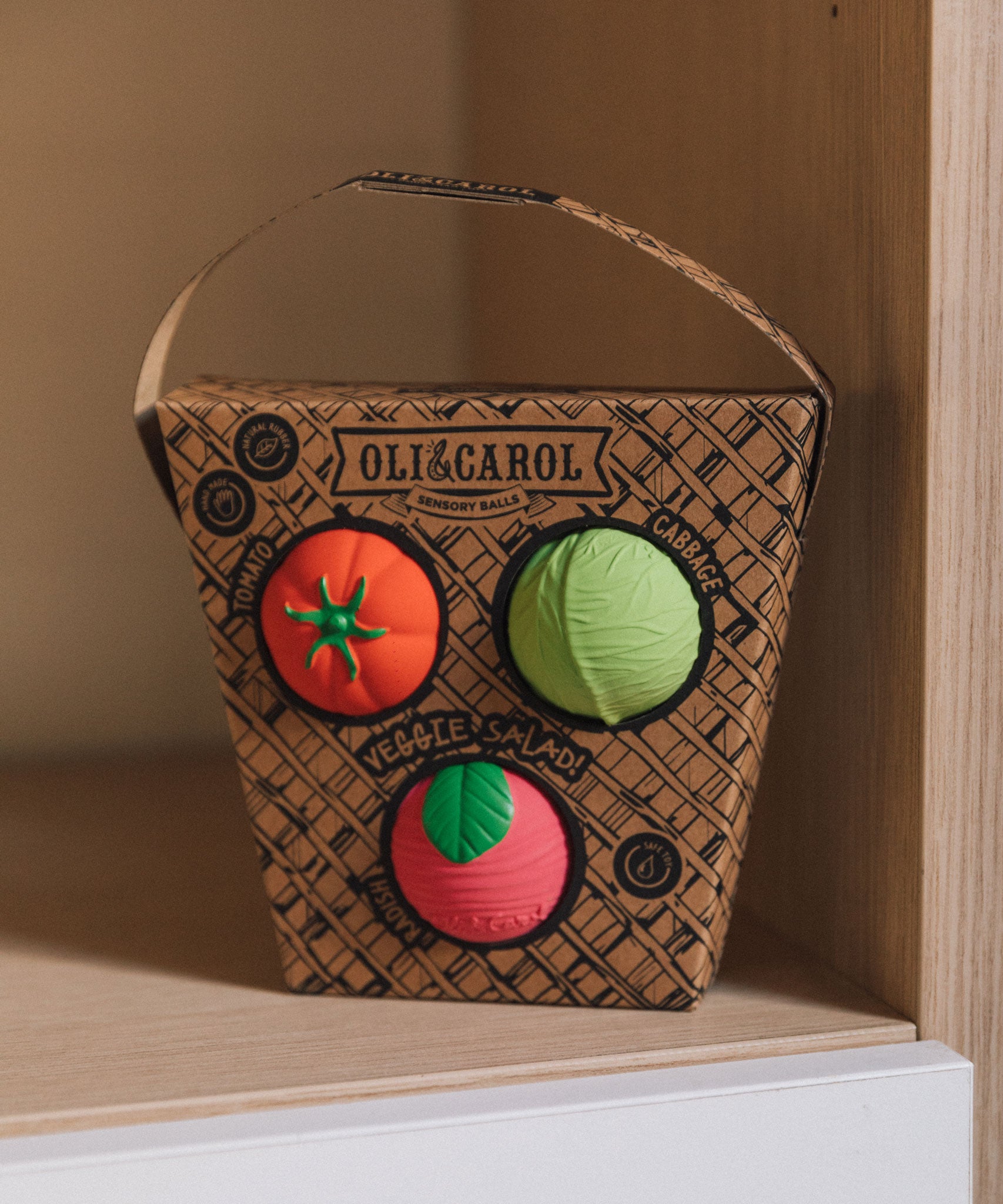 Oli & Carol Veggie Salad Baby Sensory Balls in their basket shaped cardboard packaging, a tomato, cabbage and a radish, placed on shelving
